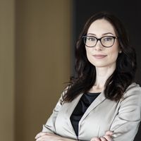 Portraitaufnahme einer professionellen Geschäftsfrau mit dunklen Haaren und Brille, gekleidet in einem hellen Blazer über einem schwarzen Top, posierend vor einem neutralen Hintergrund.