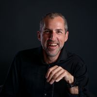 Ein Porträt eines lachenden Mannes in schwarzem Hemd vor einem dunklen Hintergrund.