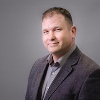 Ein Porträt eines lächelnden Mannes in einem grauen Anzug vor einem grauen Hintergrund.