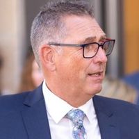 Ein Mann mittleren Alters trägt einen blauen Anzug und eine Brille. Er steht im Freien und scheint in ein Gespräch vertieft zu sein.