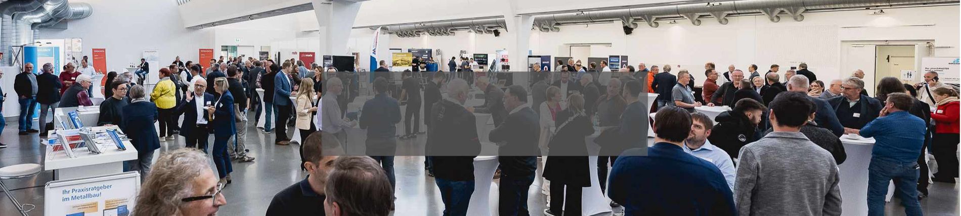 Eine große Messehalle mit vielen Besuchern, die sich um Ausstellungsstände versammeln und in Gesprächen vertieft sind.