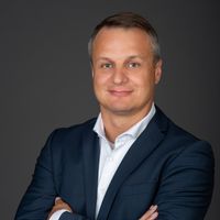 Ein Mann in einem dunklen Anzug posiert mit verschränkten Armen vor einem neutralen Hintergrund.