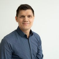 Ein Mann in einem dunkelblauen Hemd lächelt freundlich in die Kamera vor einem neutralen Hintergrund.