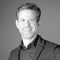 Ein Porträt eines lächelnden Mannes in einem schwarzen Hemd und Sakko vor einem grauen Hintergrund.