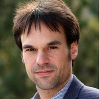 Ein Mann in einem blauen Sakko und kariertem Hemd steht draußen vor einem verschwommenen Hintergrund.