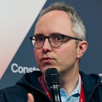 Eine Person mit Brille hält ein Mikrofon und spricht auf einer Veranstaltung.