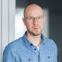 Ein Mann mit Brille und Jeanshemd steht vor einem Fenster.