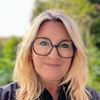 Ein Porträt einer Frau mit blonden Haaren und Brille, die in die Kamera lächelt. Der Hintergrund ist unscharf und grün.