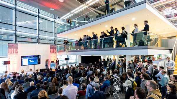 Eine große Konferenzveranstaltung mit zahlreichen Teilnehmern, die einer Präsentation auf einer Bühne folgen. Die Veranstaltung findet in einem modernen Gebäude mit zwei Ebenen statt.