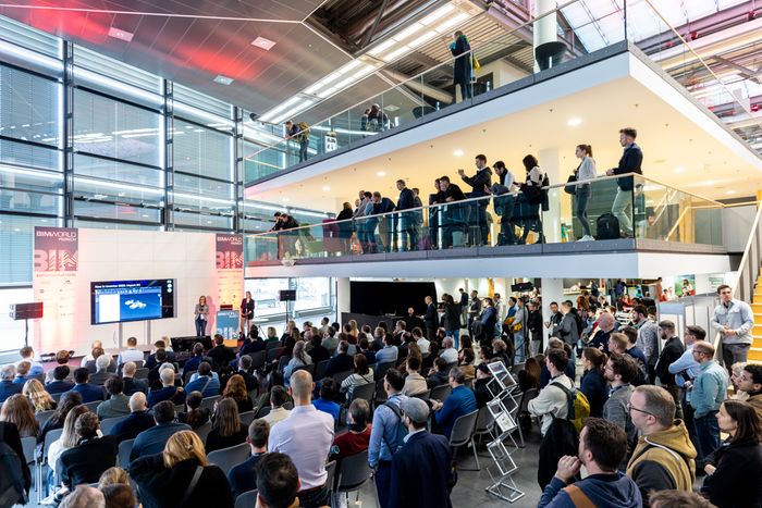 Eine große Konferenzveranstaltung mit zahlreichen Teilnehmern, die einer Präsentation auf einer Bühne folgen. Die Veranstaltung findet in einem modernen Gebäude mit zwei Ebenen statt.