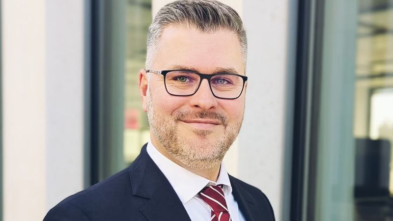 Porträt von Johannes Gaide in dunklem Anzug und gestreifter Krawatte vor einem modernen Bürogebäude.