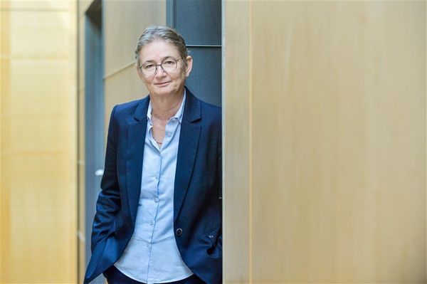 Elke Herbst in einem blauen Anzug steht in einem modernen Flur und lehnt sich an die Wand.