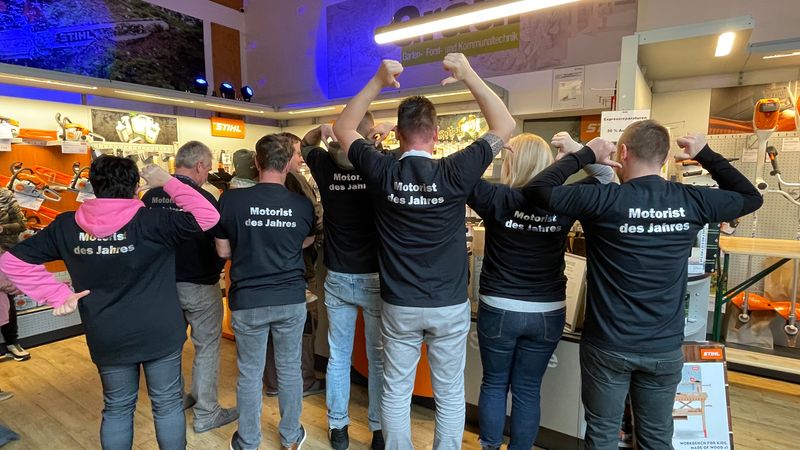 Eine Gruppe von Menschen steht in einem Geschäft und trägt schwarze T-Shirts mit der Aufschrift "Motorist des Jahres".