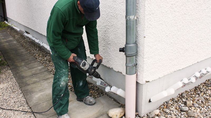 Handwerker bohrt an der Hausfassade: Ein Mann verwendet einen Bohrhammer, um Löcher nahe dem Fundament neben einem Fallrohr in die Außenwand zu bohren; er trägt Arbeitskleidung und steht auf einem Kiesweg.