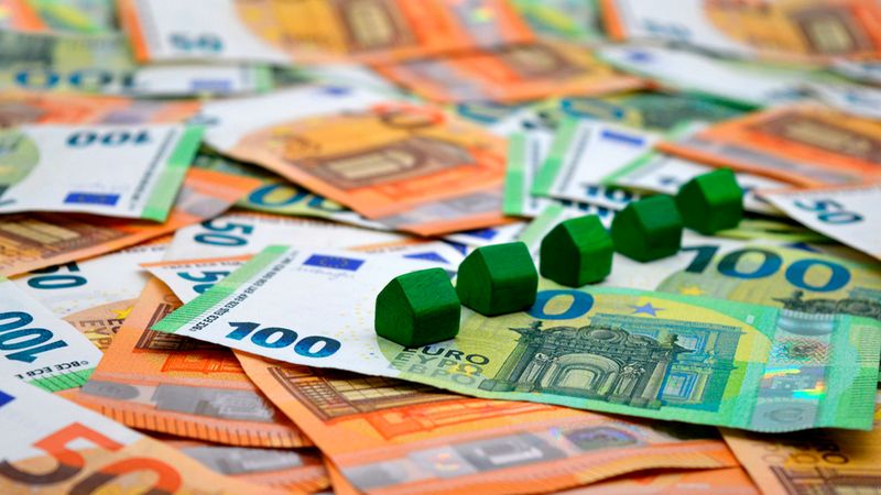 Eine Reihe kleiner grüner Holzhäuser, die auf einem verstreuten Haufen von Euro-Banknoten, darunter 50- und 100-Euro-Scheine, platziert sind und das Konzept einer Immobilieninvestition symbolisieren.