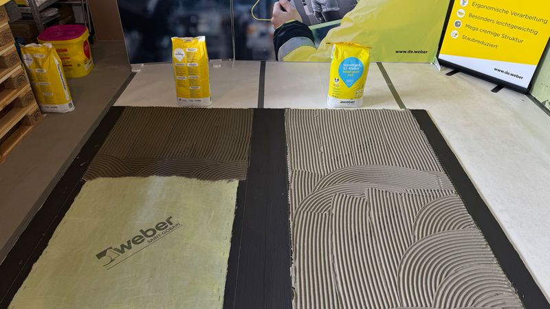 Ein Vergleich von zwei verschiedenen Fliesenklebern auf dem Boden, aufgetragen mit einer Zahnkelle. Im Hintergrund sind Säcke von weber.maxit sowie ein Informationsaufsteller mit Produktvorteilen zu sehen.