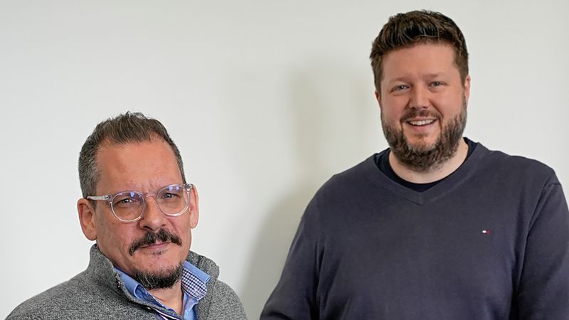 Zwei Männer stehen neben einem Regal mit Werkzeugen und Büchern.