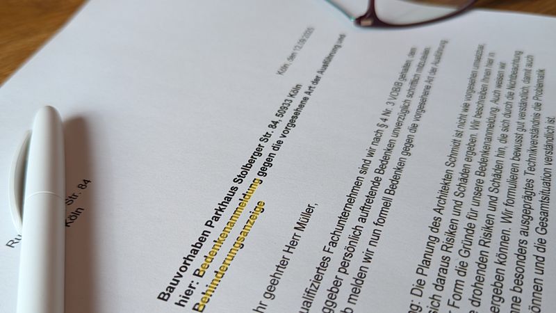 Beispielhafte Darstellung einer Bedenkenanzeige. Ein Dokument mit dem Titel "Bedenkenanzeige" liegt auf einem Tisch, daneben ein weißer Stift und eine Brille.