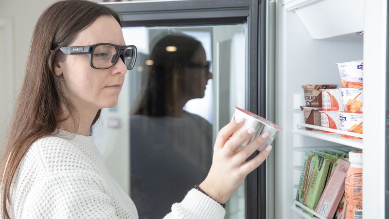 Eine Frau steht vor einem Kühlschrank und hält einen Joghurtbecher in der Hand.