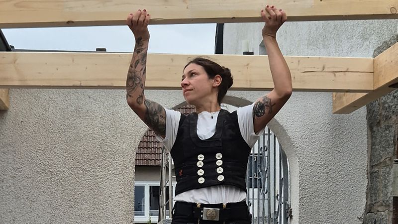 Ein Zimmermann steht auf einer Leiter und arbeitet an einem Holzbalkenrahmen.