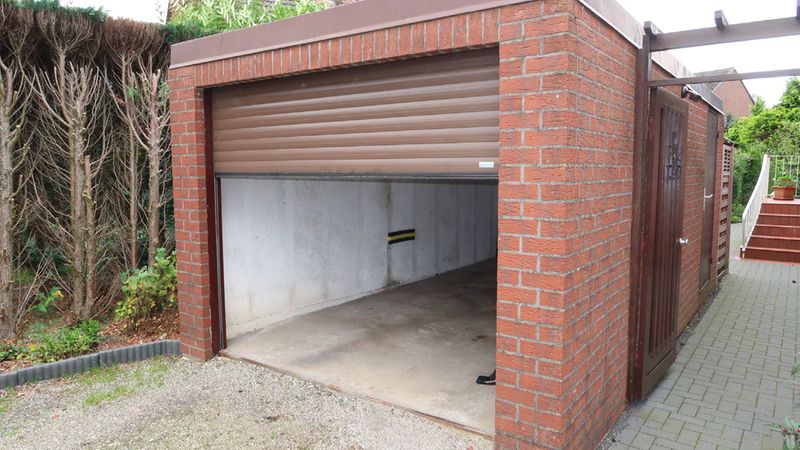 Eine offene Garage aus Backstein mit einem hochgezogenen Rolltor, umgeben von Hecken und einem gepflasterten Weg.