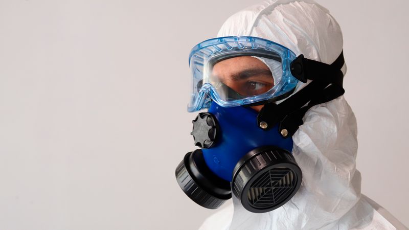 Hauptmotiv: Eine Person trägt einen weißen Schutzanzug, eine blaue Filter-Atemschutzmaske und eine transparente Schutzbrille. Die Nahaufnahme zeigt die Filterpatronen, Befestigungsriemen und die Abdichtung der Maske vor einem neutralen Hintergrund. Geeignet zur Darstellung von persönlicher Schutzausrüstung, Arbeitssicherheit, Labor- oder Gefahrstoffschutz.