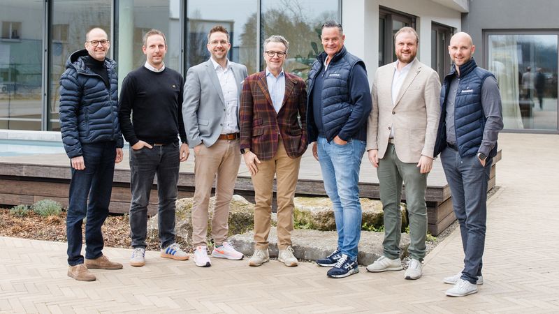 Eine Gruppe von sieben Männern steht vor einem modernen Gebäude. Sie tragen verschiedene Freizeit- und Businesskleidung und lächeln in die Kamera.