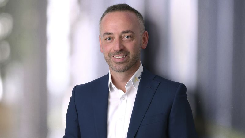 Ein Mann in einem dunkelblauen Anzug lehnt lächelnd an einer Wand in einem modernen Bürogebäude.