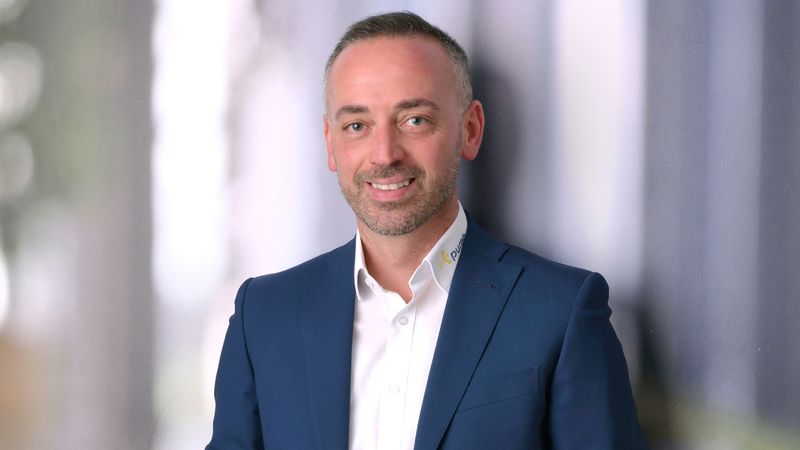 Ein Mann in einem dunkelblauen Anzug lehnt lächelnd an einer Wand in einem modernen Bürogebäude.