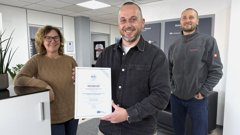 Drei Personen in einem Büro, eine Person hält ein Zertifikat in der Hand.