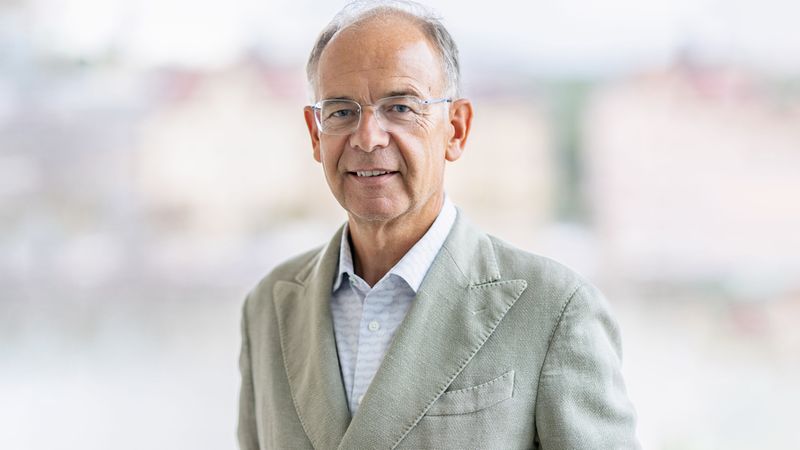 Ein älterer Mann in einem grauen Anzug steht vor einem verschwommenen Hintergrund und lächelt in die Kamera.