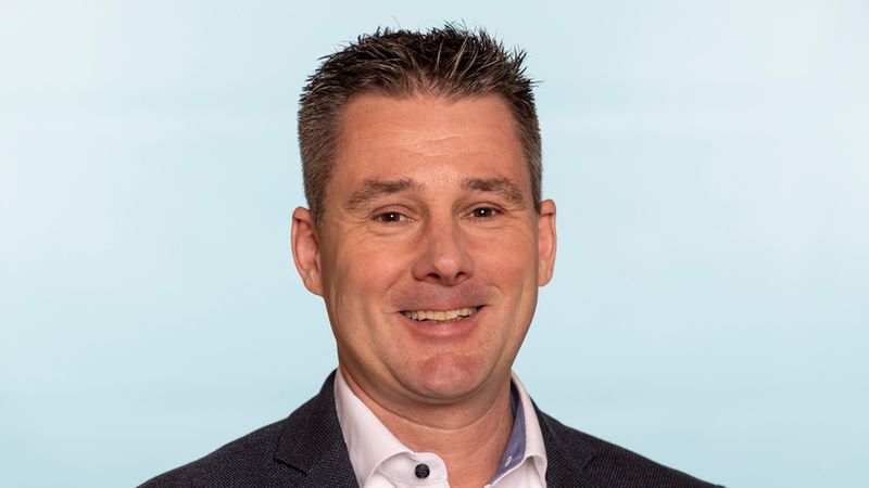 Ein Mann in einem dunklen Anzug mit weißem Hemd steht vor einem hellblauen Hintergrund.