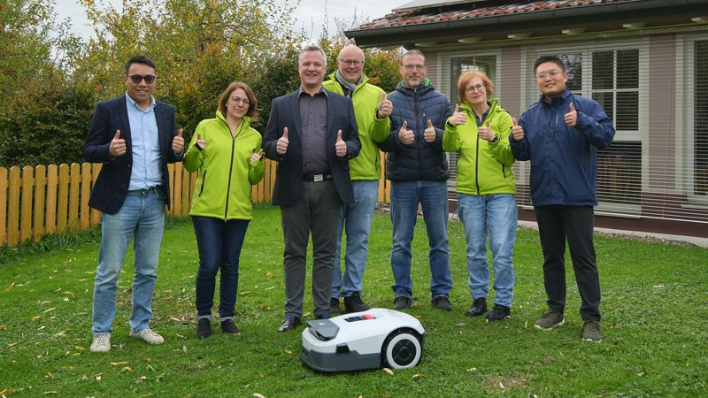 Eine Gruppe von Menschen steht im Freien und zeigt Daumen hoch neben einem Rasenroboter.