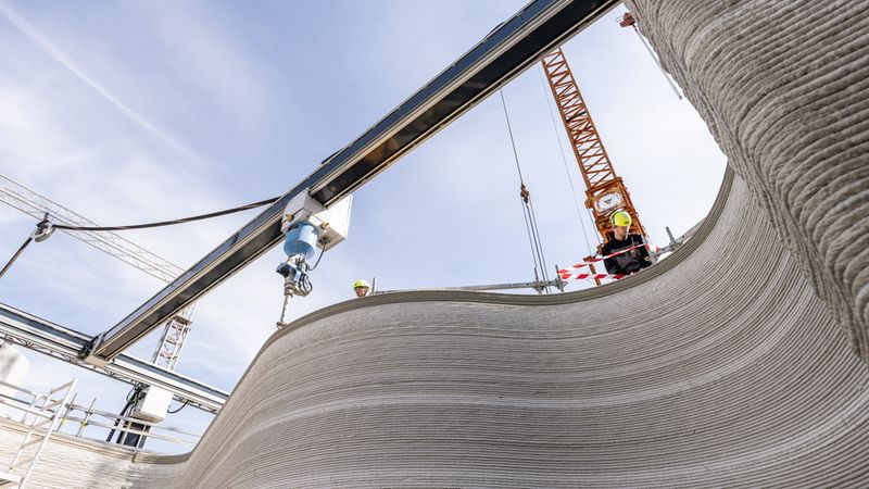 Ein 3D-Drucker erstellt die Wände eines Gebäudes auf einer Baustelle, während ein Kran im Hintergrund sichtbar ist.