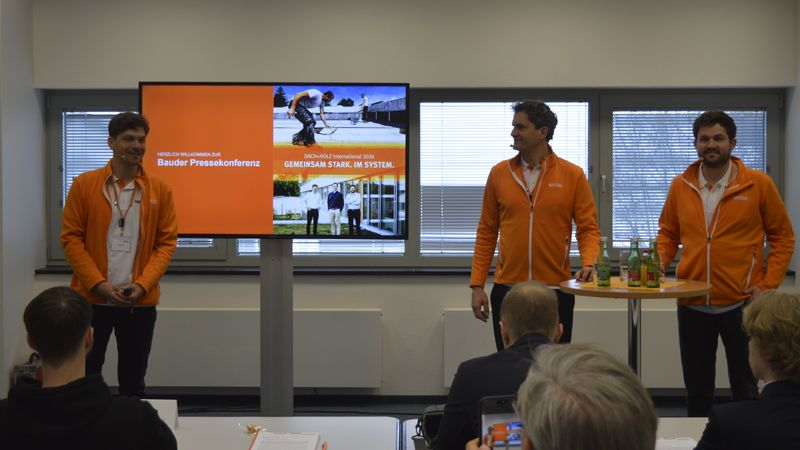 Drei Männer in orangefarbenen Jacken präsentieren bei einer Bauder Pressekonferenz vor einem Bildschirm.