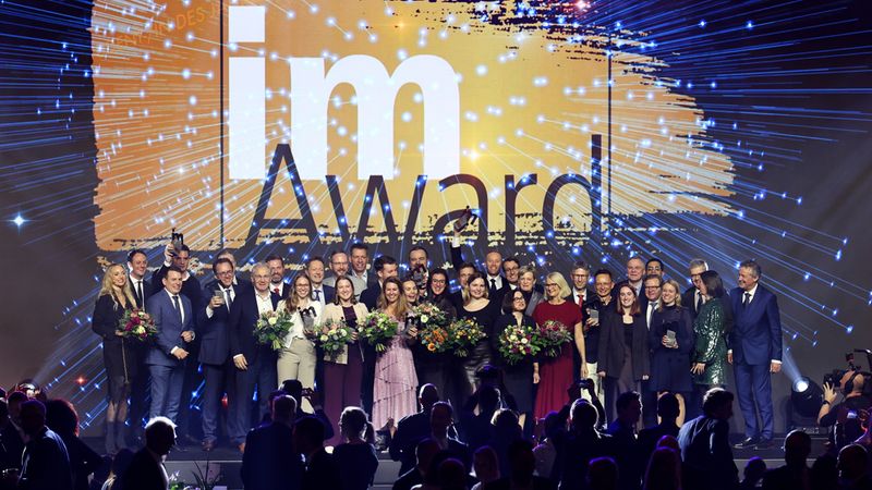 Eine große Gruppe von Menschen posiert auf einer Bühne während einer Preisverleihung. Im Hintergrund ist ein großes Schild mit der Aufschrift "Award" zu sehen.