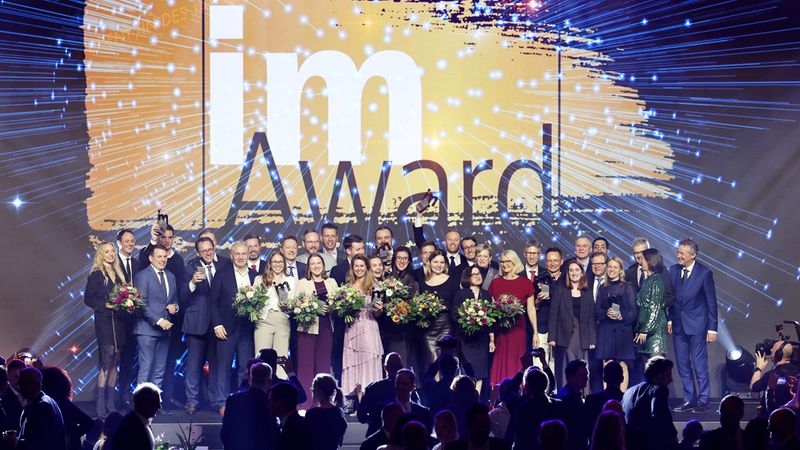 Eine große Gruppe von Menschen posiert auf einer Bühne während einer Preisverleihung. Im Hintergrund ist ein großes Schild mit der Aufschrift "Award" zu sehen.