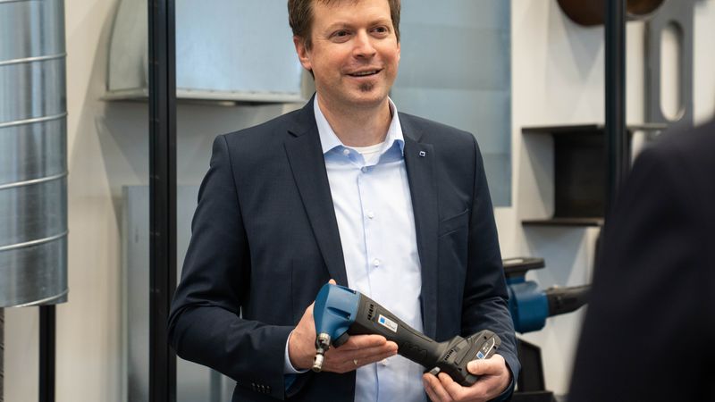 Ein Mann im Anzug hält ein Elektrowerkzeug in der Hand und lächelt in einer industriellen Umgebung.