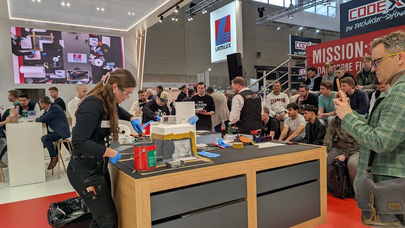 Eine Handwerksmesse mit einer Vorführung, bei der eine Frau an einem Tisch arbeitet, während Zuschauer zuschauen.