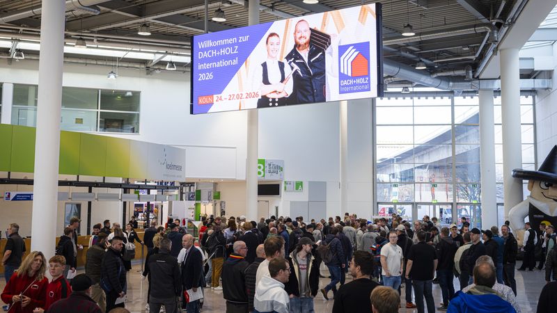 Besucher in einer Messehalle mit einem großen Bildschirm, der die DACH+HOLZ International 2026 ankündigt.