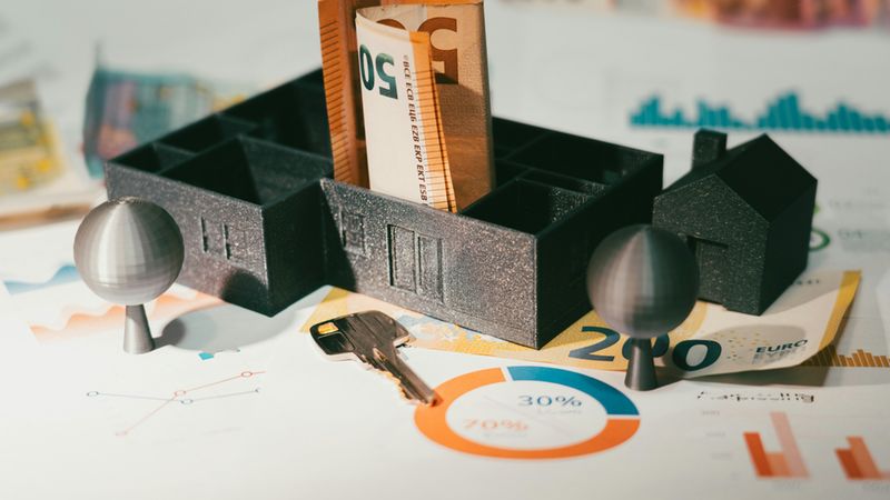 Ein Hausmodell mit Geldscheinen und einem Schlüssel auf Finanzdokumenten.