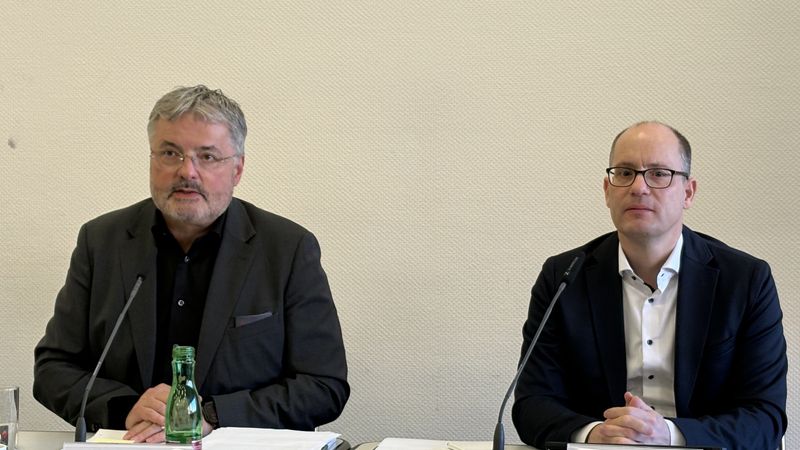 Zwei Männer sitzen an einem Tisch mit Mikrofonen und sprechen.