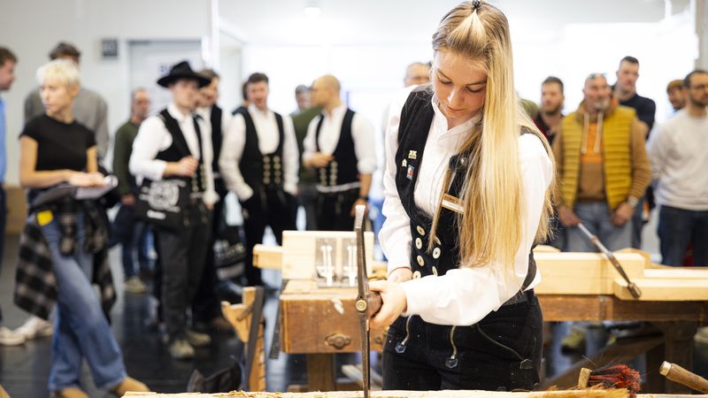 Eine Handwerkerin in traditioneller Zunftkleidung arbeitet konzentriert an einem Holzstück, während eine Gruppe von Menschen im Hintergrund zuschaut.