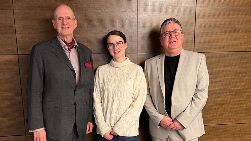 Eine Gruppe von drei Personen steht vor einer Holzwand. Die Personen tragen unterschiedliche Kleidung, darunter Anzug und Freizeitkleidung.