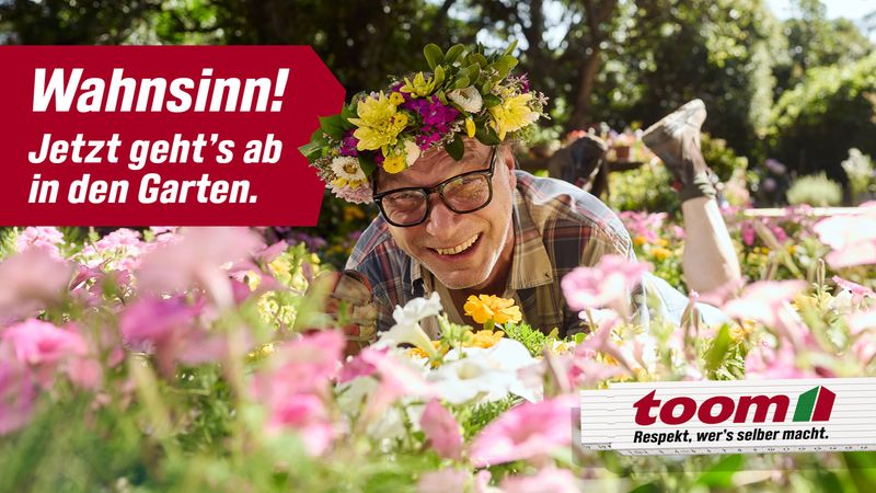 Ein Mann mit Brille und Blumenkranz liegt lächelnd in einem blühenden Garten.