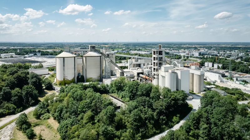 Ein großes Zementwerk umgeben von grüner Landschaft und Bäumen, mit einem weiten Blick auf die Umgebung und den Himmel.