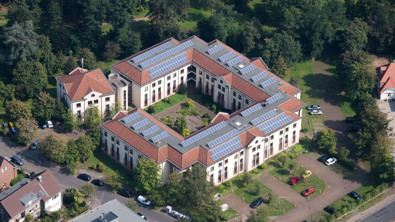 Eine Luftaufnahme eines großen Gebäudekomplexes mit Solarpaneelen auf dem Dach, umgeben von Bäumen und Grünflächen.