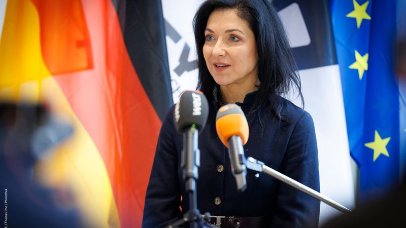 Eine Frau steht vor mehreren Mikrofonen und spricht, im Hintergrund sind die Flaggen von Deutschland und der EU zu sehen.