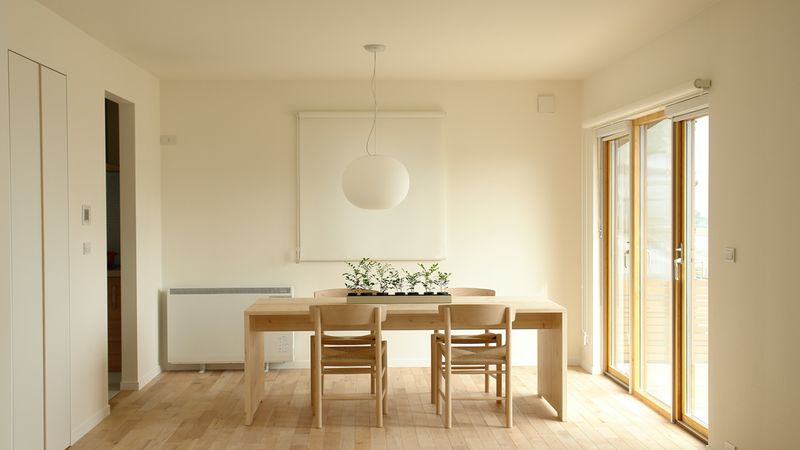 Ein minimalistisches Esszimmer mit einem Holztisch, vier Stühlen und einer hängenden Lampe, das von natürlichem Licht durch große Fenster erhellt wird.