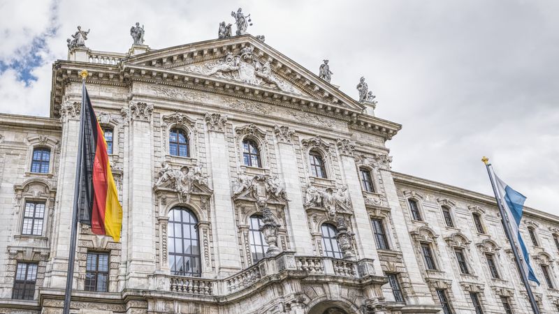 Der Justizpalast in München mit seiner beeindruckenden neobarocken Fassade und den deutschen sowie bayerischen Flaggen im Vordergrund.